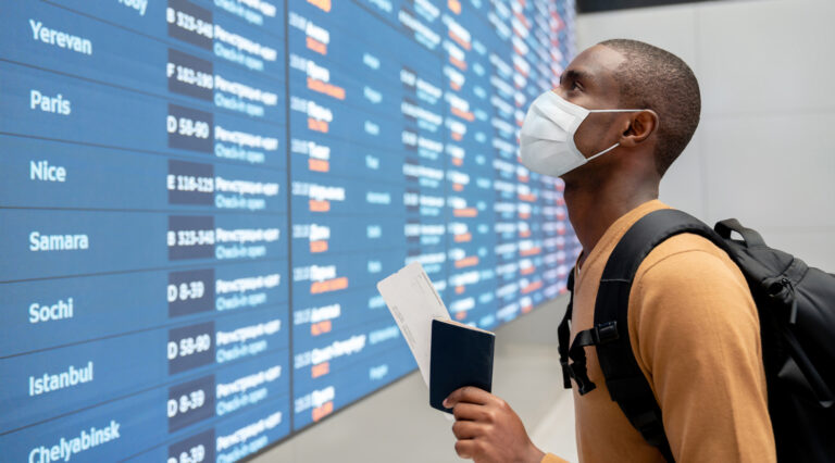 Portrait of a black male traveler wearing a facemask at the airport and looking at the flight schedule - travel concepts