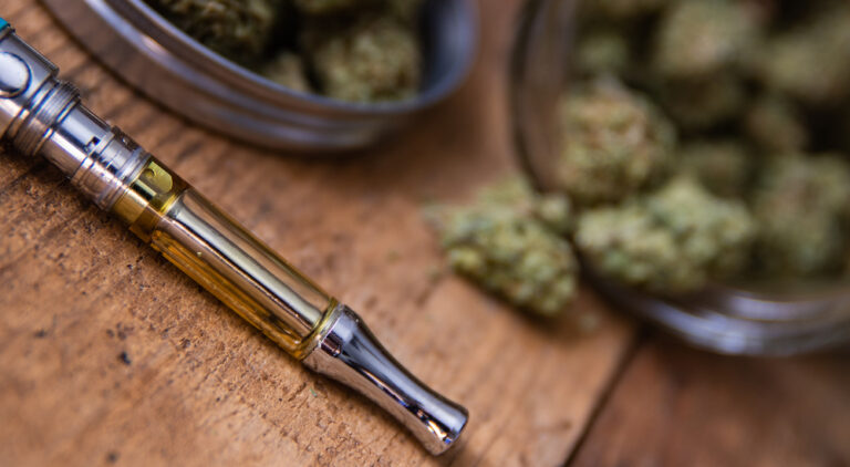 THC oil concentrate filled vape pen on natural wood with an open glass container full of Mango Kush strain marijuana buds grown &amp; sold in dispensaries through-out Southern California.