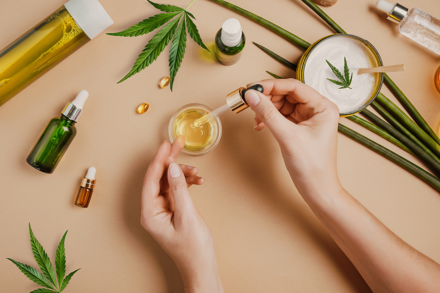 Pipette with CBD cosmetic oil in female hands on a table background with cosmetics, cream with cannabis and hemp leaves, marijuana. Flat lay, top view.