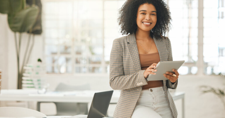A picture of a woman in a suite jacket sitting in an office. She has a tablet in her hand, and she's smiling.