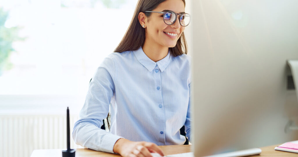 Smiling female payment service provider using mouse, stylus and tablet next to her on wooden desk in small office