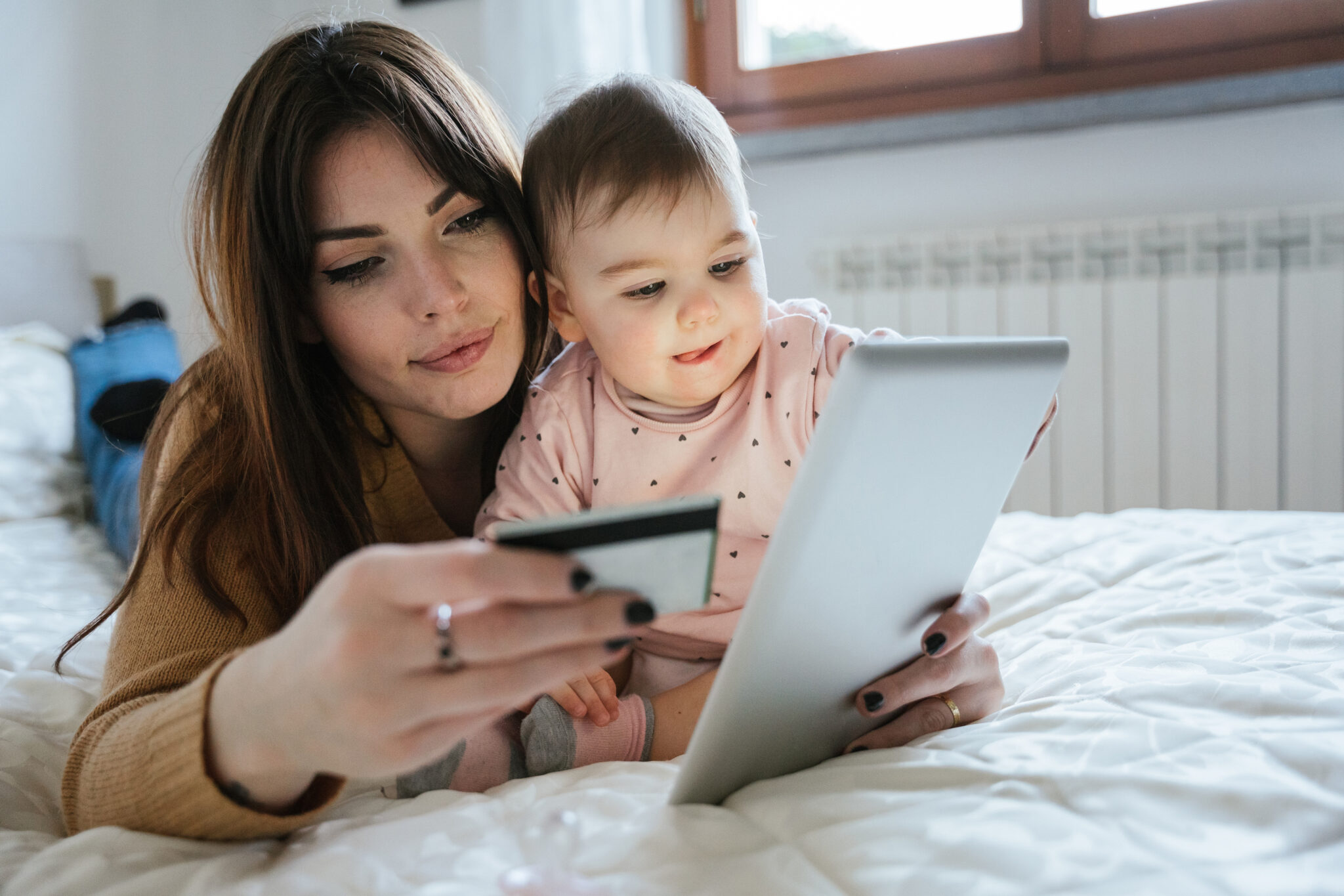 Portrait of a beautiful young mom with a baby girl lying on the bed in a moment of intimacy while buying products online with the tablet and holding her credit card in hand - Millennial with daughter Portrait of a beautiful young mom with a baby girl lying on the bed in a moment of intimacy while buying products online with the tablet and holding her credit card in hand - Millennial with daughter