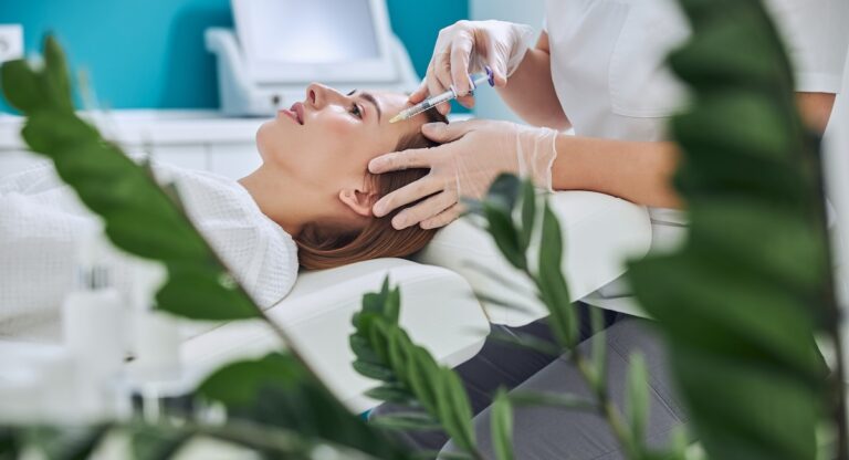Waist up side view portrait of elegant redheaded woman lying on the medicine chair while beautician taking face injection with special product in beauty center
