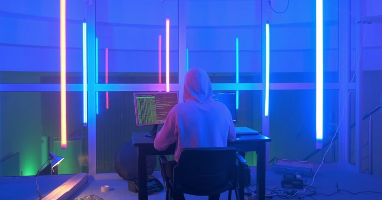 Back view of criminal hacker working in the evening on computer in big office with warm colored neon lights. Computer terrorism.