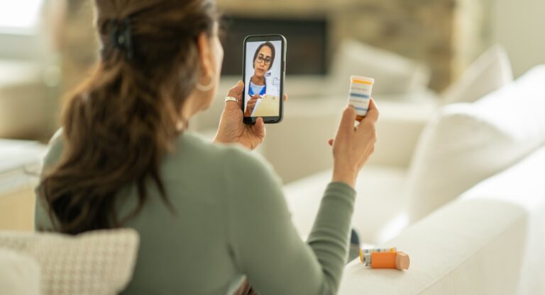 A senior woman of Middle Eastern decent sits on the sofa in the comfort of her own home as she meets with her doctor virtually.  She has her medications in hand as she video chats her doctor on her cell phone.