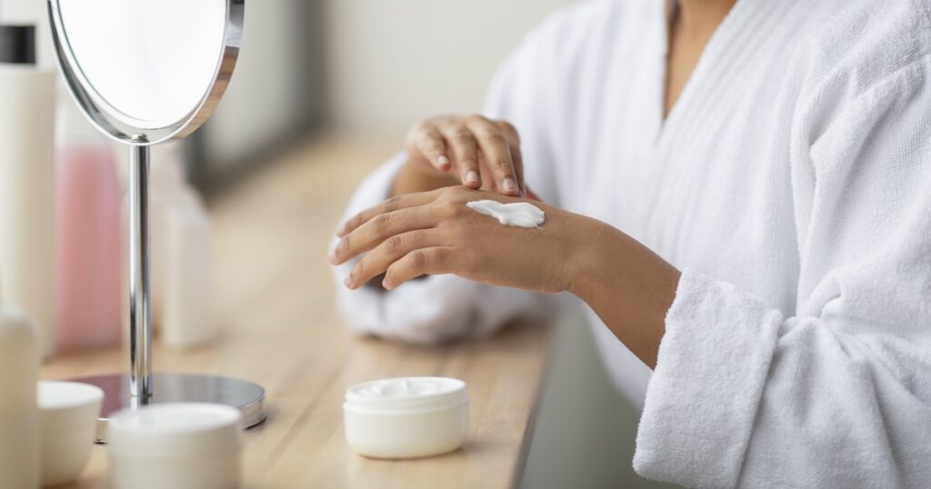 Cropped of indian woman enjoying her morning body care routine Cropped of young indian woman wearing white bathrobe sitting next to window, enjoying her morning body care routine, applying cream moisturizer on her hand after shower, bathroom interior