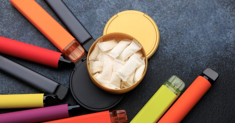 Nicotine pouches are inside a round container while several electronic cigarettes are arranged around it. The surface is dark and flat. The colors of the products vary.