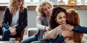 Caring female counselor hugs a female patient during a group therapy session.