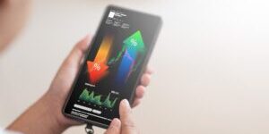 A close-up shot shows a person's hand holding a smartphone, displaying a complex financial analysis interface with upward and downward trending percentage arrows, stock market charts, and various data points. This image is perfect for illustrating concepts like investment, trading, financial growth, economic analysis, and mobile financial management.