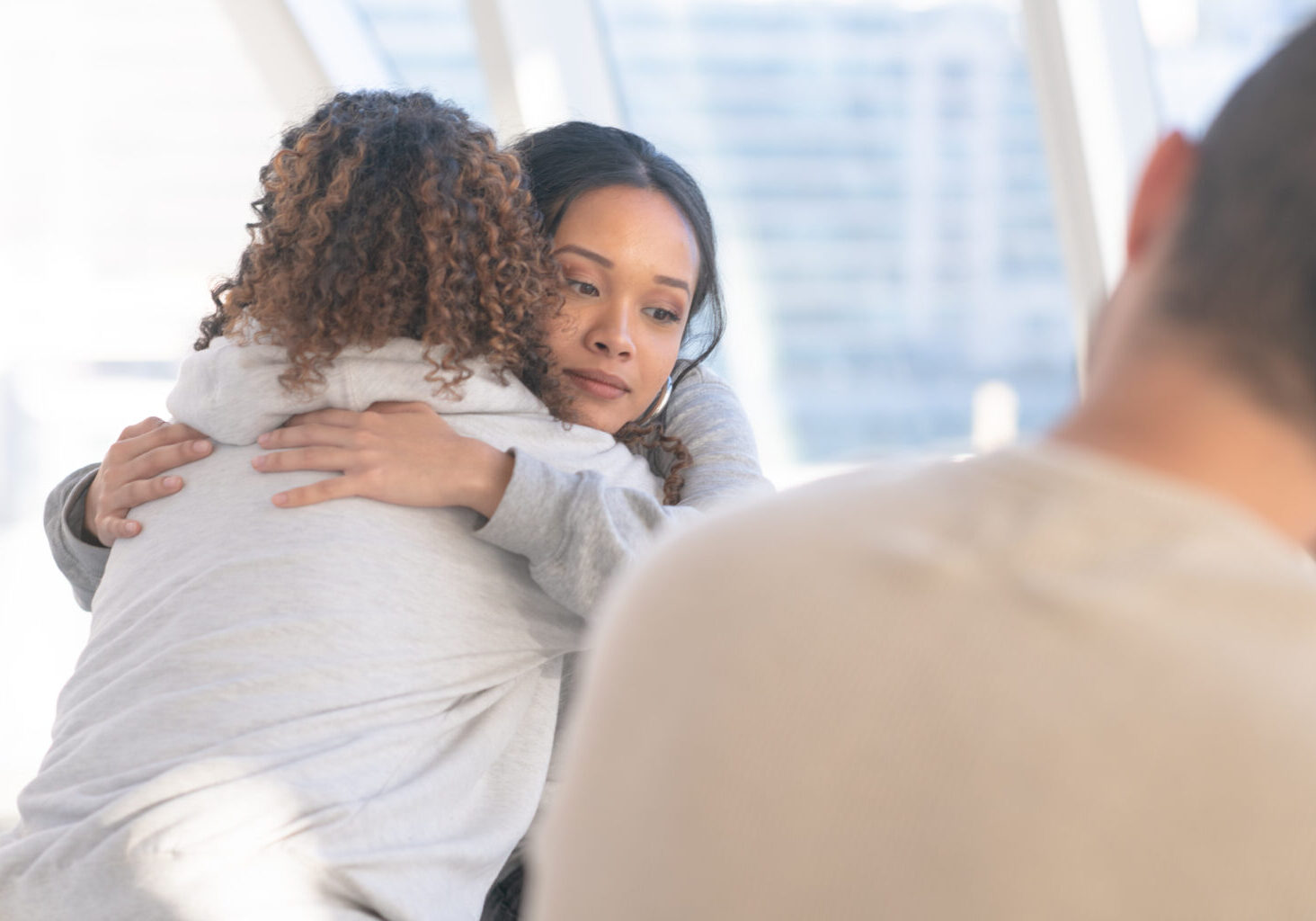 Embracing in group therapy A group of young and diverse adults sits in a circle of chairs one afternoon. They are having a group therapy session regarding their addiction to recreational drugs. Two (2) women embrace in a comforting hug.