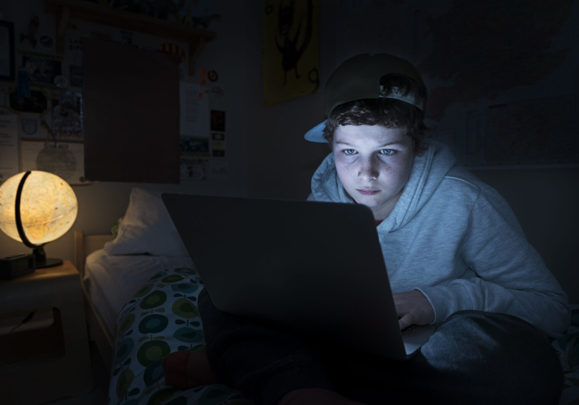 Portrait of a young teenage boy online in his bedroom. He is lying on his bed in a darkened room and he is illuminated by the light coming from the screen from his laptop as he surfs the web late at night.