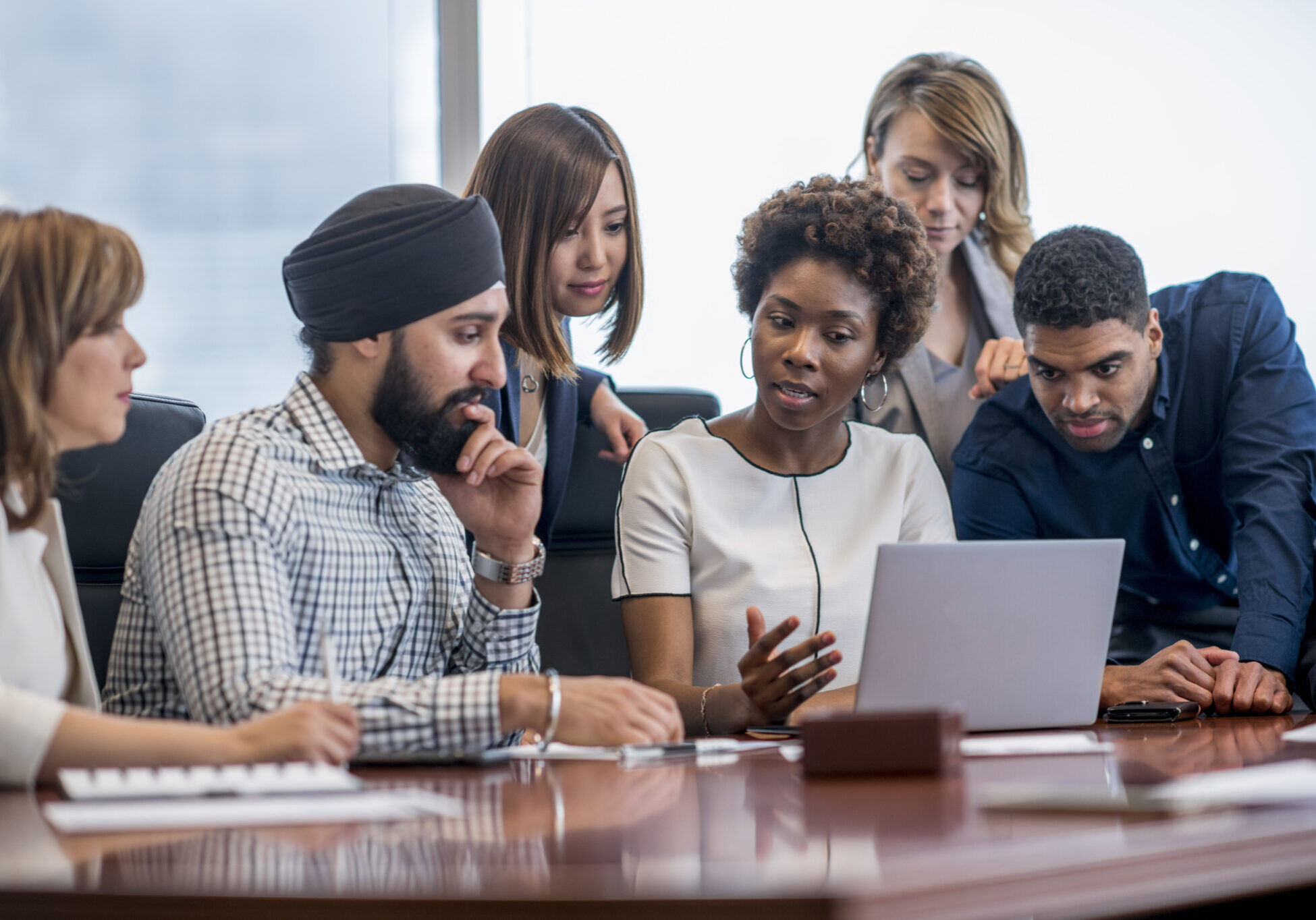 A diverse group of business people gather around a laptop in a modern office and discuss what they see.