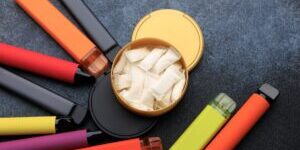 Nicotine pouches are inside a round container while several electronic cigarettes are arranged around it. The surface is dark and flat. The colors of the products vary.