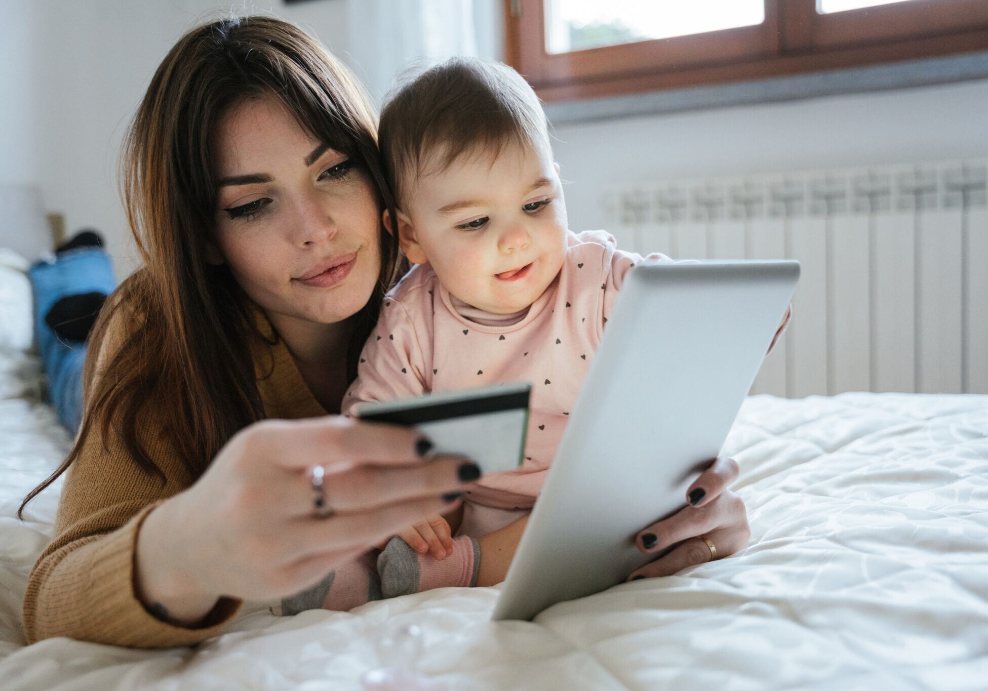 Portrait of a beautiful young mom with a baby girl lying on the bed in a moment of intimacy while buying products online with the tablet and holding her credit card in hand - Millennial with daughter